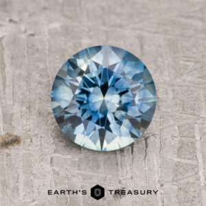 A 1.37-Carat Teal Blue Montana Sapphire (Heated), round and faceted, sparkles on a textured gray surface with the "Earth's Treasury" logo and text at the bottom.