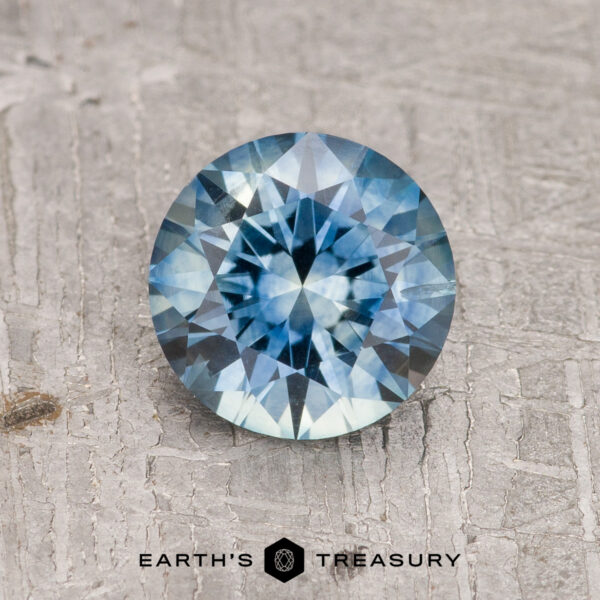 1.37-Carat Teal Blue Montana Sapphire (Heated) A 1.37-Carat Teal Blue Montana Sapphire (Heated), round and faceted, sparkles on a textured gray surface with the "Earth's Treasury" logo and text at the bottom.