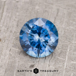 A 1.27-carat medium blue heated Montana Sapphire, round brilliant-cut, sits on a textured gray surface with "EARTH'S TREASURY" displayed below.