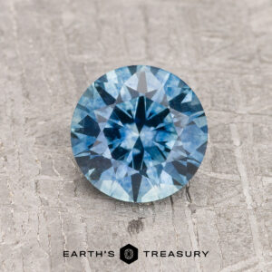 A 1.13-carat Teal Blue Montana Sapphire (Heated), round brilliant-cut, is shown on a gray textured surface with "EARTH'S TREASURY" displayed below.