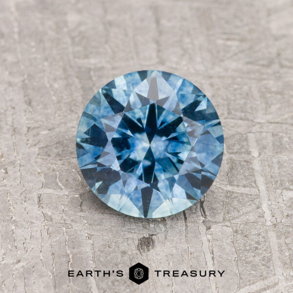 1.13-Carat Teal Blue Montana Sapphire (Heated) A 1.13-carat Teal Blue Montana Sapphire (Heated), round brilliant-cut, is shown on a gray textured surface with "EARTH'S TREASURY" displayed below.