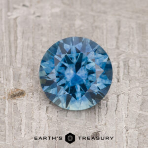 A 1.19-carat rich robin's egg blue Montana Sapphire (heated), round and faceted, sits on a textured gray surface with the "Earth's Treasury" logo displayed at the bottom.
