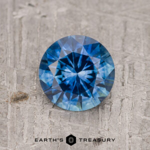 A 1.14-carat rich blue Montana Sapphire (heated), round brilliant-cut, is shown on a textured gray surface with "EARTH'S TREASURY" visible beneath the gem.