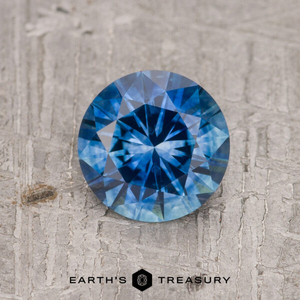1.14-Carat Rich Blue Montana Sapphire (Heated) A 1.14-carat rich blue Montana Sapphire (heated), round brilliant-cut, is shown on a textured gray surface with "EARTH'S TREASURY" visible beneath the gem.