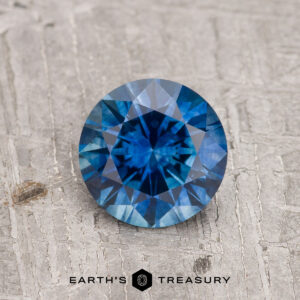 1.06-Carat Rich Blue Montana Sapphire (Heated) in round brilliant cut displayed on a textured gray surface, featuring the "Earth's Treasury" logo at the bottom.