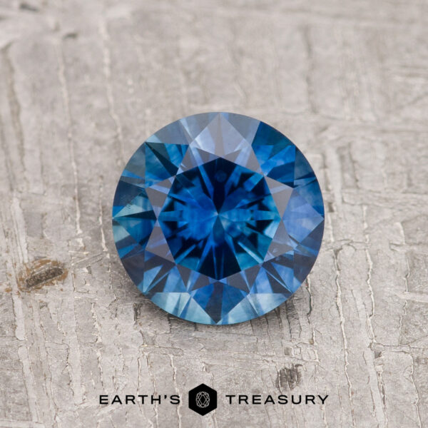 1.06-Carat Rich Blue Montana Sapphire (Heated) 1.06-Carat Rich Blue Montana Sapphire (Heated) in round brilliant cut displayed on a textured gray surface, featuring the "Earth's Treasury" logo at the bottom.