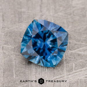A 1.76-carat medium blue heated Montana Sapphire with a cushion cut rests on a textured gray surface, with the "Earth's Treasury" logo and name displayed at the bottom.