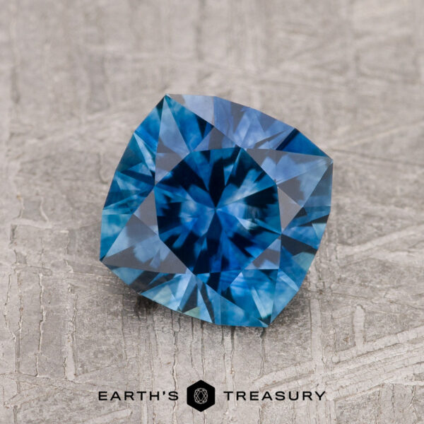 1.76-Carat Medium Blue Montana Sapphire (Heated) A 1.76-carat medium blue heated Montana Sapphire with a cushion cut rests on a textured gray surface, with the "Earth's Treasury" logo and name displayed at the bottom.