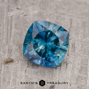 The 1.56-Carat Peacock Blue Montana Sapphire (Heated), cushion-cut, rests elegantly on a textured gray surface, with the Earth’s Treasury logo and text beneath.