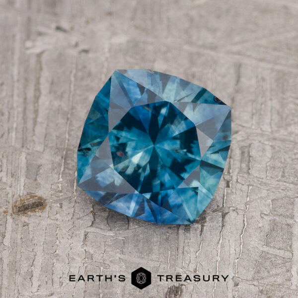 1.56-Carat Peacock Blue Montana Sapphire (Heated) The 1.56-Carat Peacock Blue Montana Sapphire (Heated), cushion-cut, rests elegantly on a textured gray surface, with the Earth’s Treasury logo and text beneath.