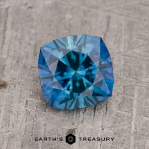 A 1.46-carat rich blue heated Montana Sapphire with a geometric cut rests on a textured gray background, "EARTH'S TREASURY" printed underneath.