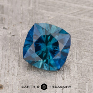 A 1.28-carat Peacock Blue Montana Sapphire (Heated) with a cushion cut rests on a textured gray surface; "EARTH'S TREASURY" appears at the bottom.