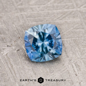 A 1.02-carat Rich Sky Blue Montana Sapphire (Heated) with a cushion cut rests on a textured gray surface, the "Earth's Treasury" logo displayed below.