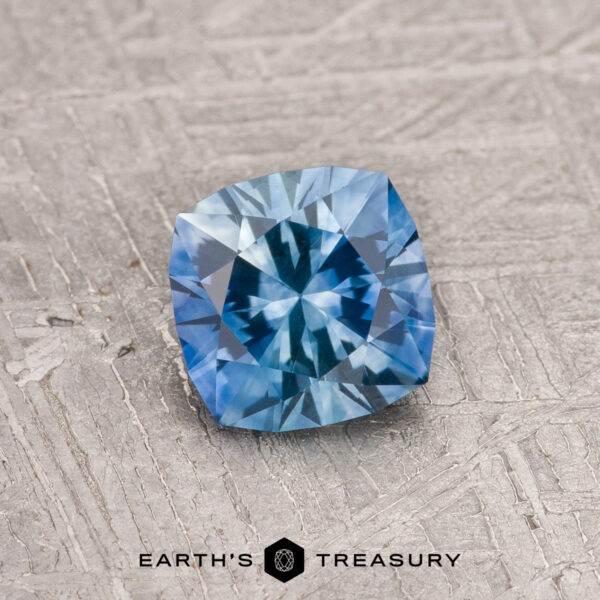1.02-Carat Rich Sky Blue Montana Sapphire (Heated) A 1.02-carat Rich Sky Blue Montana Sapphire (Heated) with a cushion cut rests on a textured gray surface, the "Earth's Treasury" logo displayed below.