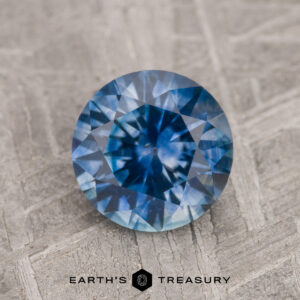 A 1.73-carat medium blue heated Montana Sapphire is showcased on a textured gray surface, featuring the "Earth's Treasury" logo at the bottom.