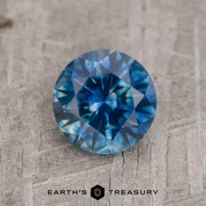 A 1.29-carat medium blue heated Montana Sapphire, round brilliant-cut, rests on a textured wooden surface with the Earth’s Treasury logo below.