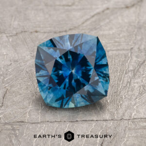 A 1.34-Carat Peacock Blue Montana Sapphire (Heated), cushion-cut with geometric facets, rests on a textured gray surface. The Earth’s Treasury logo is shown at the bottom of the image.