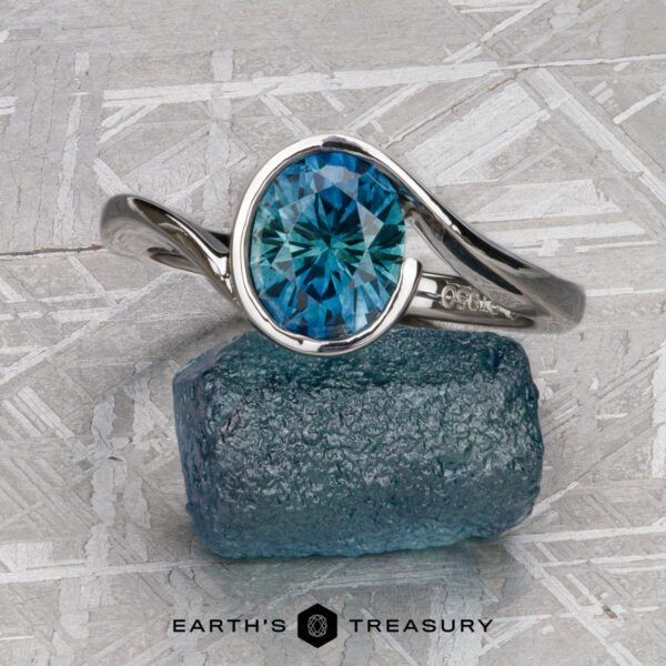 The "Calliste" ring in platinum with 1.66-Carat Montana Sapphire A platinum "Calliste" ring featuring a 1.66-carat Montana Sapphire is displayed on a rough blue stone, set against a geometric patterned background with the text "EARTH'S TREASURY" at the bottom.