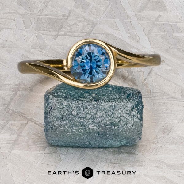 The "Calliste" ring in 18K yellow gold with 1.08-carat Montana sapphire The "Calliste" ring in 18K yellow gold with a 1.08-carat Montana sapphire is displayed on a rough blue stone, with the "EARTH'S TREASURY" logo beneath—a striking example of fine gemstone jewelry.