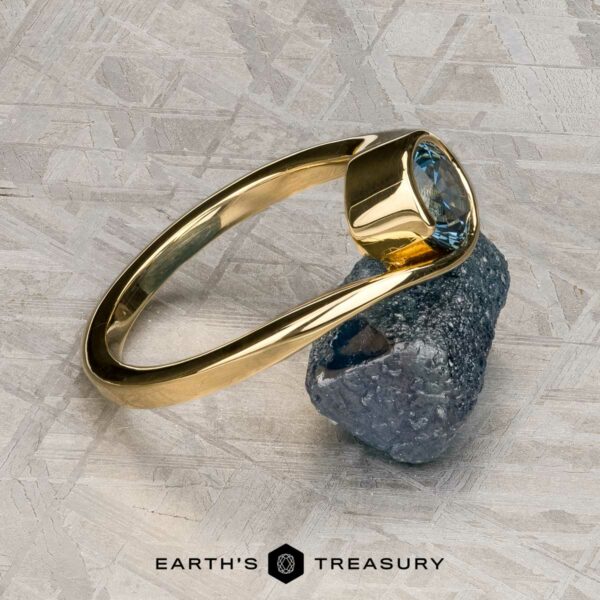 The "Calliste" ring in 18K yellow gold with 1.08-carat Montana sapphire The "Calliste" ring in 18K yellow gold, set with a 1.08-carat Montana sapphire, rests on a rough dark mineral atop a textured gray surface. The Earth’s Treasury logo is displayed below.