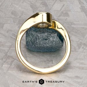 The "Calliste" ring in 18K yellow gold with a 1.08-carat Montana sapphire is shown upright on a gray textured surface, with the "Earth's Treasury" logo and text at the bottom.
