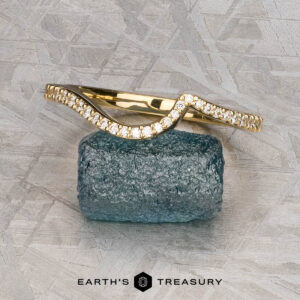 The "Calliste" band in 18K yellow gold, featuring a wavy design set with small diamonds, is displayed on a rough blue stone with the text "EARTH'S TREASURY" below.
