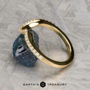 The "Calliste" band in 18K yellow gold, featuring a row of small diamonds, rests on a rough dark blue stone against a textured grey background. The text "EARTH'S TREASURY" is displayed at the bottom.