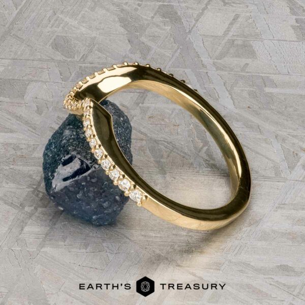 The "Calliste" band in 18K yellow gold The "Calliste" band in 18K yellow gold, featuring a row of small diamonds, rests on a rough dark blue stone against a textured grey background. The text "EARTH'S TREASURY" is displayed at the bottom.