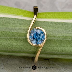 The "Calliste" ring in 18K yellow gold with a 1.08-carat Montana sapphire from Calliste Jewelry is shown on a green and white striped plant stem, with the "EARTH'S TREASURY" logo at the bottom.
