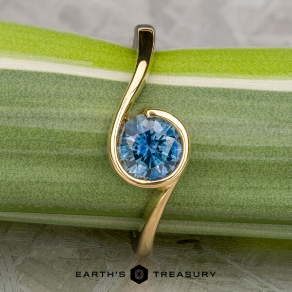 The "Calliste" ring in 18K yellow gold with 1.08-carat Montana sapphire The "Calliste" ring in 18K yellow gold with a 1.08-carat Montana sapphire from Calliste Jewelry is shown on a green and white striped plant stem, with the "EARTH'S TREASURY" logo at the bottom.