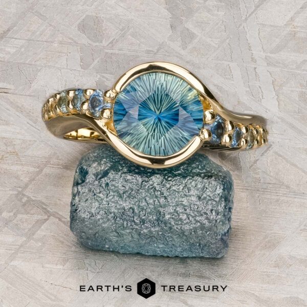 The "Venus" ring in 14K yellow gold with 2.14-carat Madagascar sapphire The "Venus" ring in 14K yellow gold, set with a 2.14-carat Madagascar sapphire and small side stones, is displayed on a rough blue mineral above the "Earth's Treasury" branding.