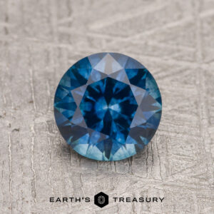 A 1.27-carat rich blue Montana Sapphire (heated), cut in a round brilliant shape, is displayed on a textured gray surface with "EARTH'S TREASURY" underneath.
