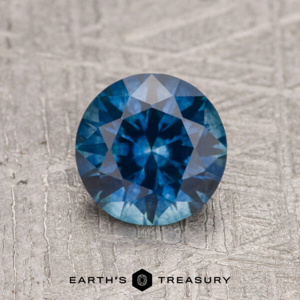 1.27-Carat Rich Blue Montana Sapphire (Heated) A 1.27-carat rich blue Montana Sapphire (heated), cut in a round brilliant shape, is displayed on a textured gray surface with "EARTH'S TREASURY" underneath.