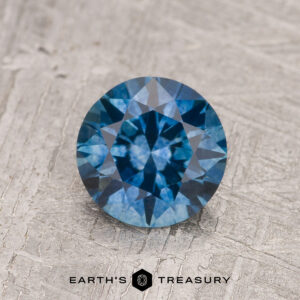 The 1.24-Carat Medium Blue Montana Sapphire (Heated) features a round brilliant cut and radiant medium blue hue, showcased on textured light gray with the "Earth's Treasury" logo beneath it.