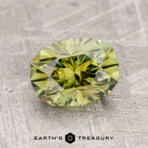 A 2.43-carat olive green demantoid garnet is displayed on a textured gray surface with the text "EARTH'S TREASURY" below.