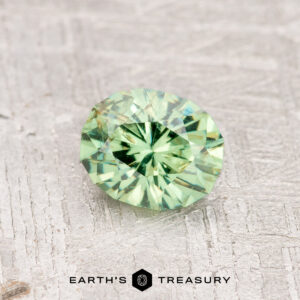 A 0.84-carat Mint Green Demantoid Garnet, expertly faceted into a round shape, rests on a textured light gray surface. The words "Earth's Treasury" appear at the bottom of the image.