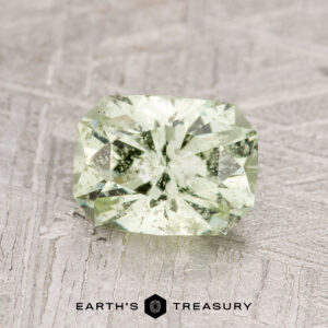 A 1.52-carat Pale Green Grossular Garnet with visible inclusions, cut in a faceted rectangular shape, is displayed on a textured surface above the "Earth's Treasury" logo.