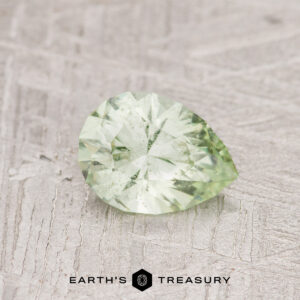 The 0.80-carat pale kiwi green grossular garnet, featuring a faceted pear shape, rests on a textured surface.