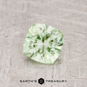 A 0.71-carat Mint Green Garnet, cushion-cut and pale green in color, rests on a textured light gray surface above the “Earth’s Treasury” logo at the bottom of the image.