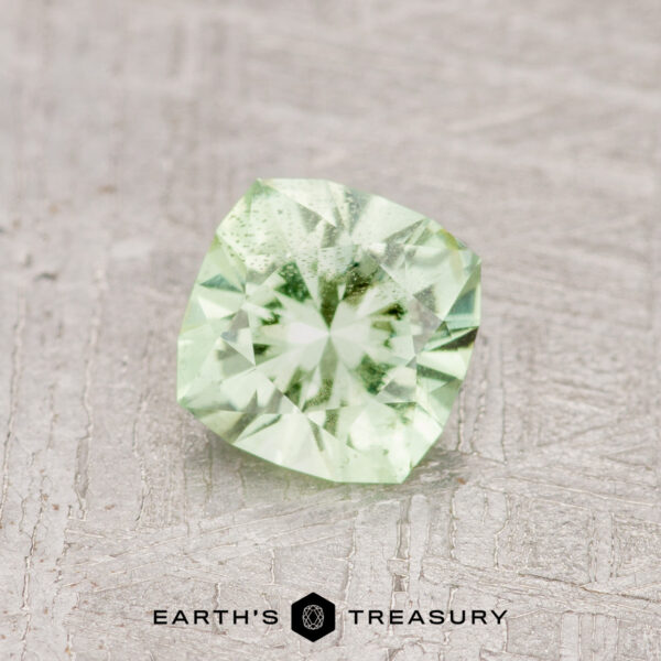 0.71-Carat Mint Green Garnet A 0.71-carat Mint Green Garnet, cushion-cut and pale green in color, rests on a textured light gray surface above the “Earth’s Treasury” logo at the bottom of the image.