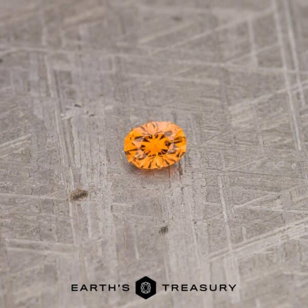 A 2.10-carat California Spessartine Garnet, small, round, and faceted with an orange hue, sits against a textured gray background. The text "EARTH'S TREASURY" is displayed at the bottom of the image.