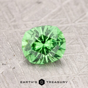 A faceted green gemstone on a textured surface, labeled "0.99-Carat Tsavorite Garnet.