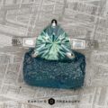 A platinum “Adelaide” ring featuring a 2.33-carat Montana sapphire rests on a rough blue stone. The background has a textured, metallic pattern. “Earth’s Treasury” is displayed at the bottom.