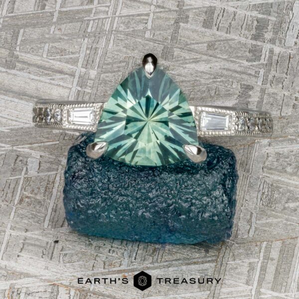 The “Adelaide” ring in platinum with 2.33-Carat Montana Sapphire A platinum “Adelaide” ring featuring a 2.33-carat Montana sapphire rests on a rough blue stone. The background has a textured, metallic pattern. “Earth’s Treasury” is displayed at the bottom.