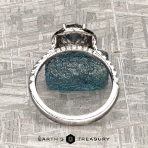 The "Victoria" Ring in 14k rose and white gold features a 1.05-carat Montana sapphire as the center stone, accented by small clear stones along the band. Displayed on a rough blue crystal with the "EARTH'S TREASURY" logo at the bottom.