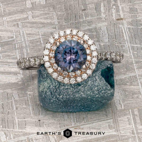 The "Victoria" Ring in 14k rose and white gold with 1.05-Carat Montana Sapphire The "Victoria" Ring in 14k rose and white gold with a 1.05-carat Montana sapphire and double diamond halo sits on a rough blue stone atop a textured metallic surface. "EARTH'S TREASURY" is displayed at the bottom.