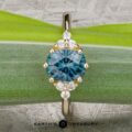 The “Mari” ring in 14k yellow gold features a 1.63-carat Montana Sapphire center, encircled by small diamonds and beautifully displayed on a green leaf background.