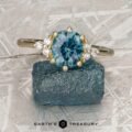 The “Mari” ring in 14k yellow gold with a 1.63-carat Montana Sapphire and small diamonds is shown on a rough blue stone, set against a textured gray background with the "Earth’s Treasury" logo.