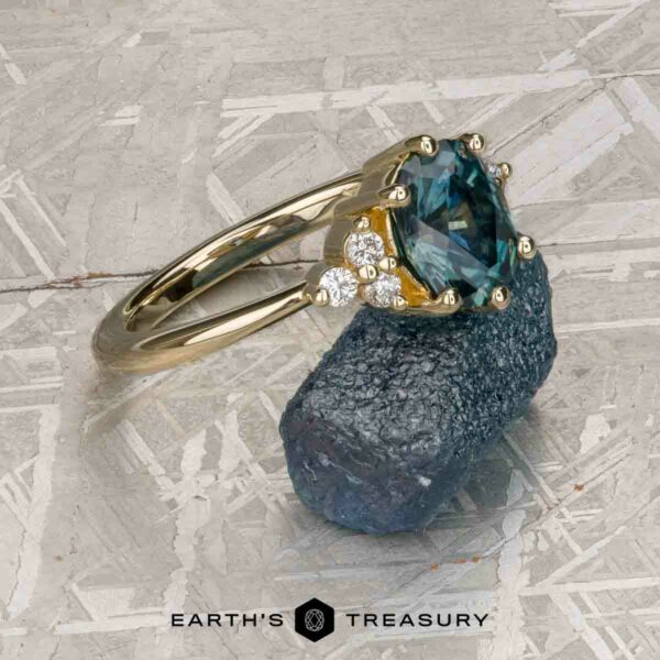 The “Mari” ring in 14k yellow gold with 1.63-Carat Montana Sapphire The “Mari” ring in 14k yellow gold with a 1.63-carat Montana sapphire and small diamonds sits on a rough blue crystal atop a patterned metallic surface. The text "Earth's Treasury" is displayed at the bottom.