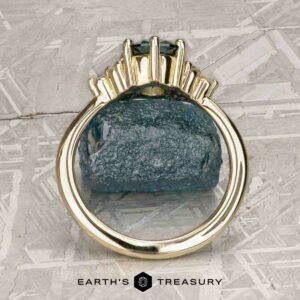 The “Mari” ring in 14k yellow gold with a 1.63-carat Montana Sapphire is shown on a rough blue stone, with the "Earth's Treasury" logo and text at the bottom.